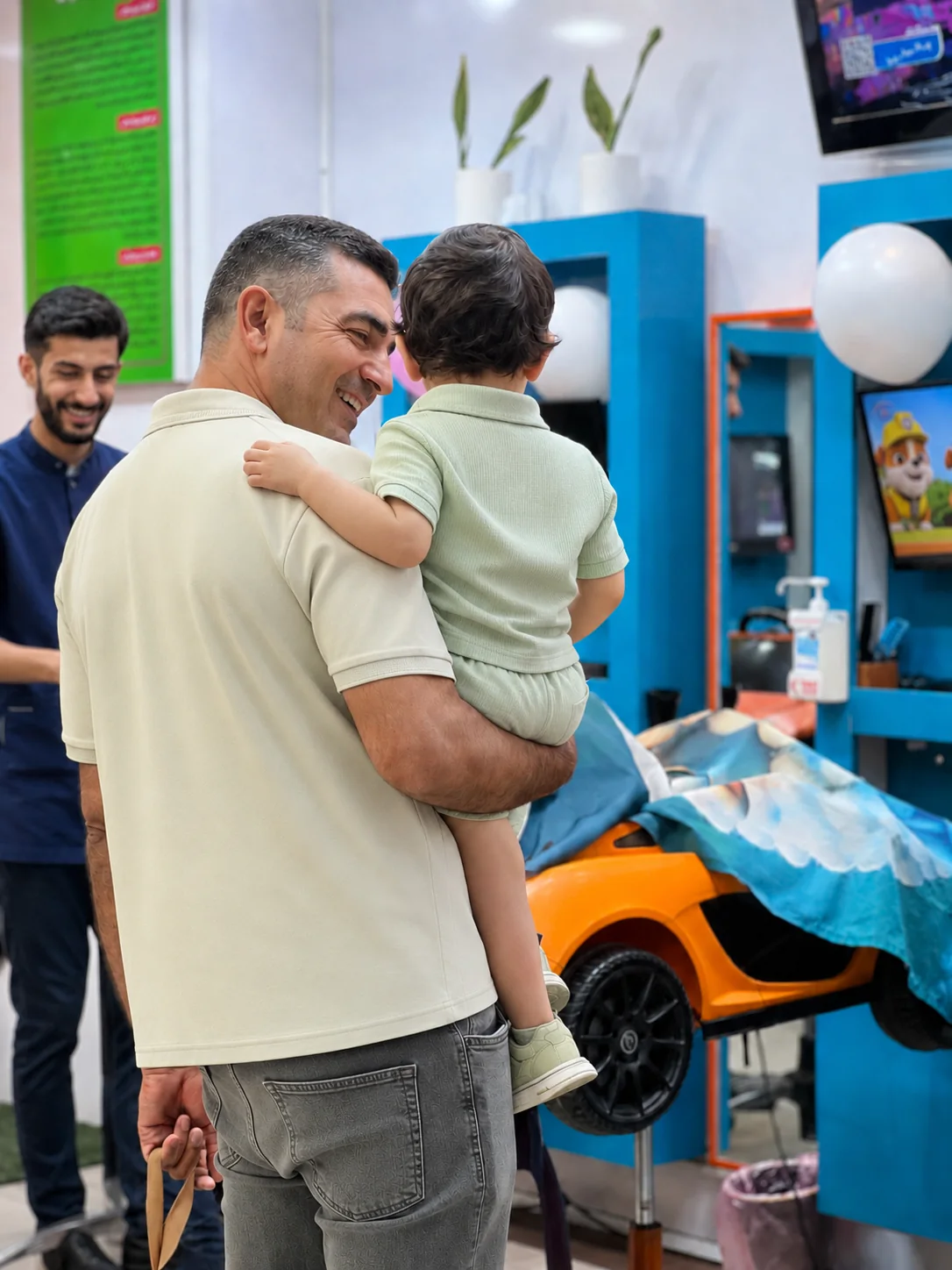 آرایشگر کودک در کنار پدر و کودک در فضای واقعی آرایشگاه کودک لبخند